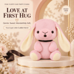 Front view of a pastel pink floppy-ear puppy plush toy wearing a black collar on a display pedestal.